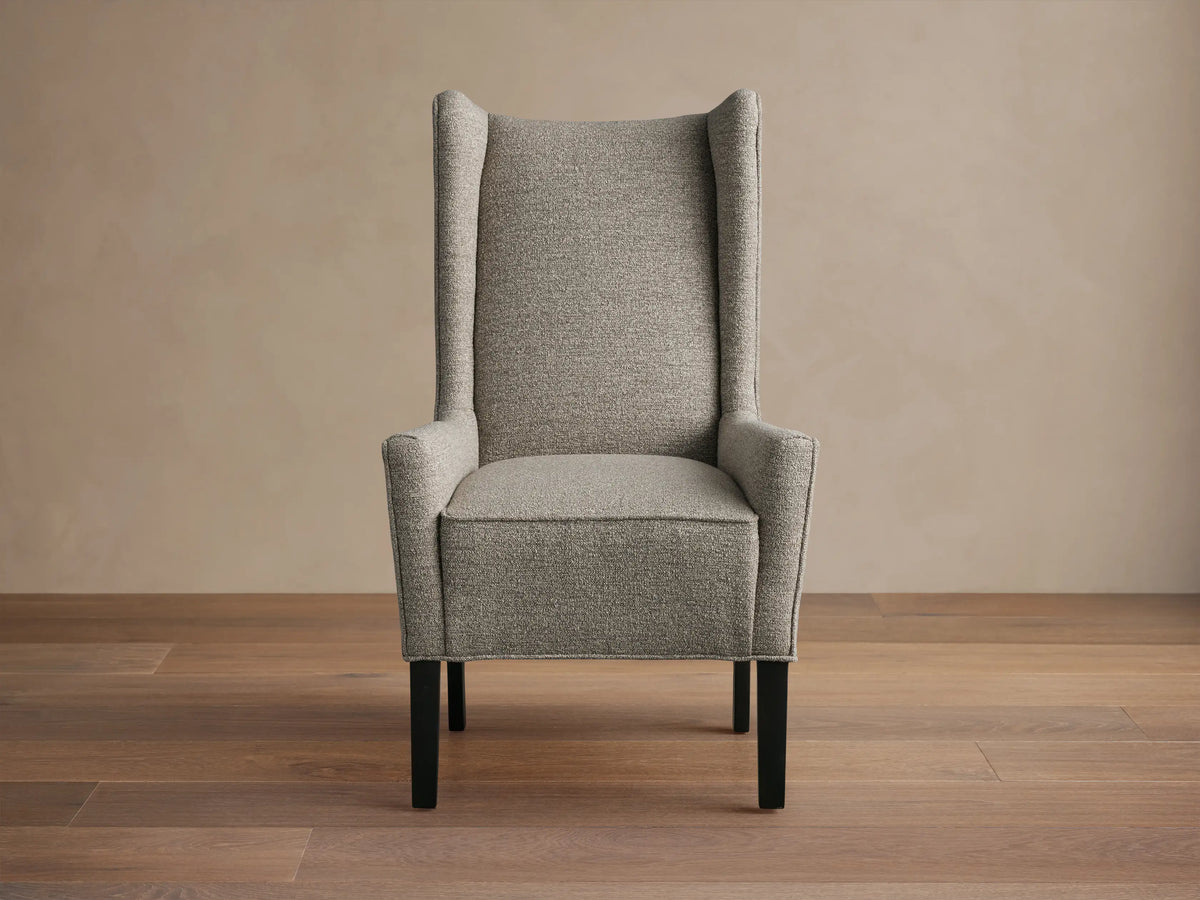 Halstead Dining Chair in gray fabric featuring a high wingback design, padded seat, and black wooden legs, set on a wooden floor.