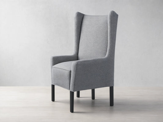 Halstead Dining Chair in gray fabric featuring a high wingback design, padded seat, and black wooden legs, set on a wooden floor.