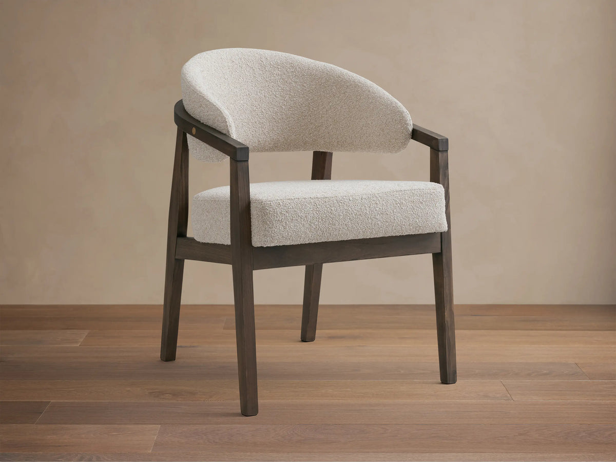 Dining chair with dark beech wood frame, curved arms with brass details, padded seat and backrest in light fabric, on wooden floor.