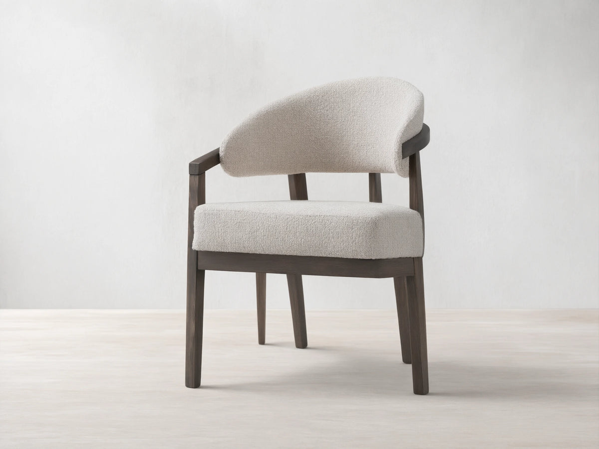 Dining chair with dark beech wood frame, curved arms with brass details, padded seat and backrest in light fabric, on wooden floor.