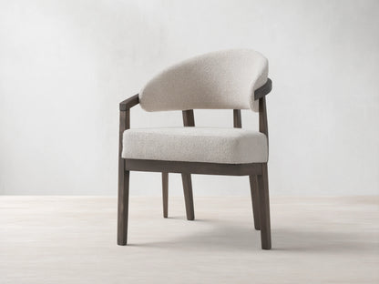 Dining chair with dark beech wood frame, curved arms with brass details, padded seat and backrest in light fabric, on wooden floor.
