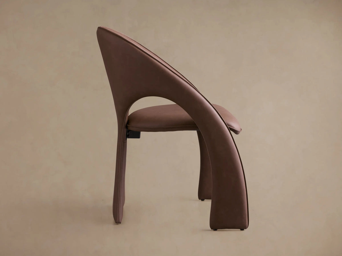Side profile of the taupe upholstered Hearst Dining Chair, with a continuous curved backrest and sculptural flared legs.