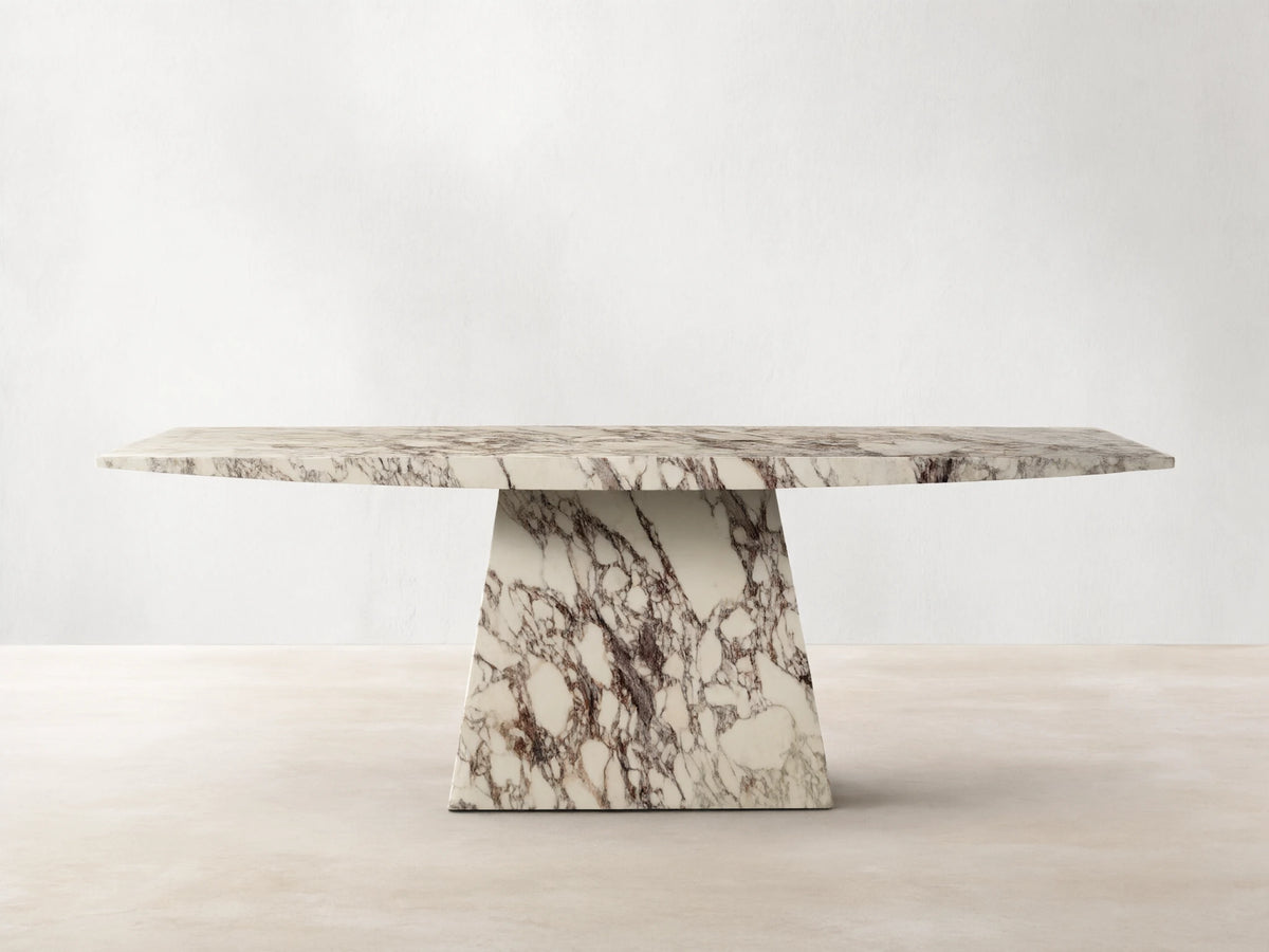 Front view of the Ilaria Dining Table, a rectangular white marble table with dark purple and brown natural veining, on a trapezoidal base.