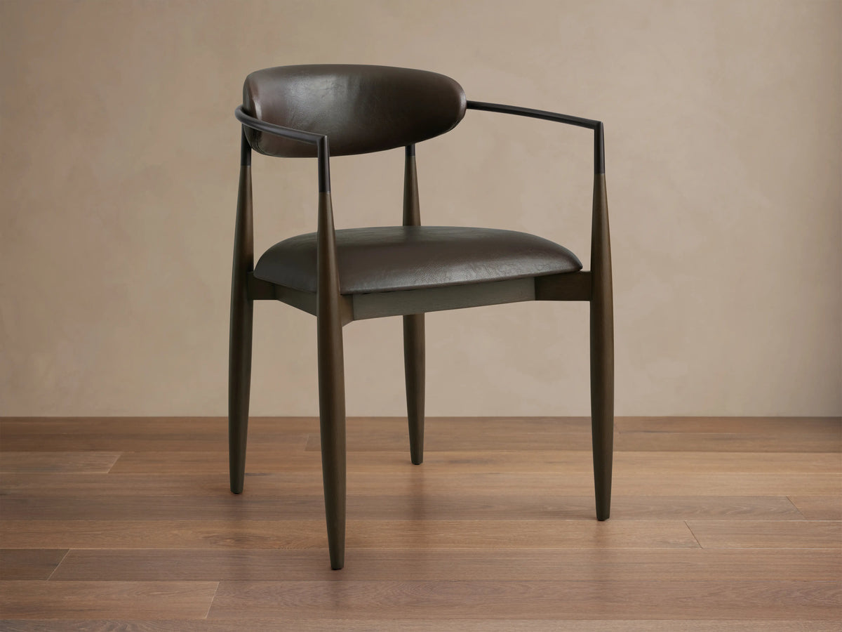 Jagger Faux Leather Dining Arm Chair in Cinder with a curved back, oil rubbed bronze accents, and tapered legs on wooden floor.