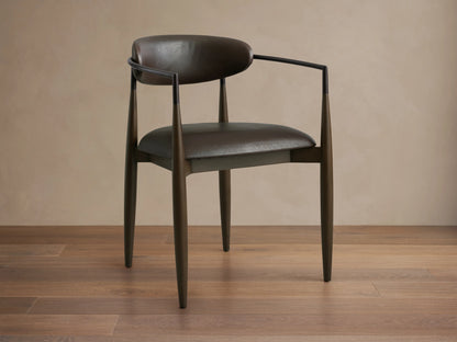 Jagger Faux Leather Dining Arm Chair in Cinder with a curved back, oil rubbed bronze accents, and tapered legs on wooden floor.