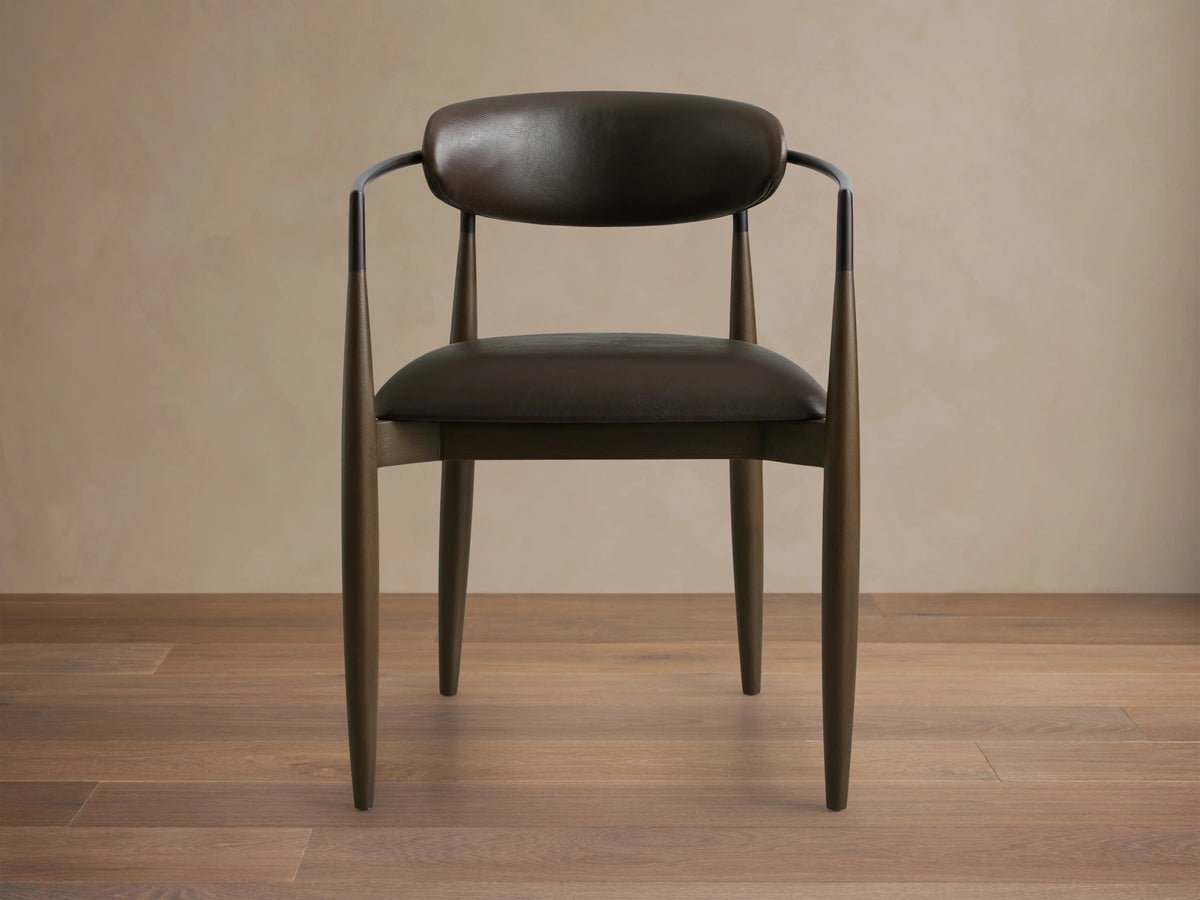 Jagger Faux Leather Dining Arm Chair in Cinder with a curved back, oil rubbed bronze accents, and tapered legs on wooden floor.