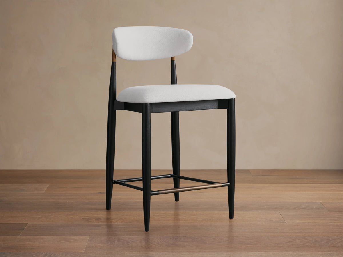 Jagger Armless Stool with a white cushioned seat and backrest, black tapered legs, and a footrest bar, on a wooden floor.