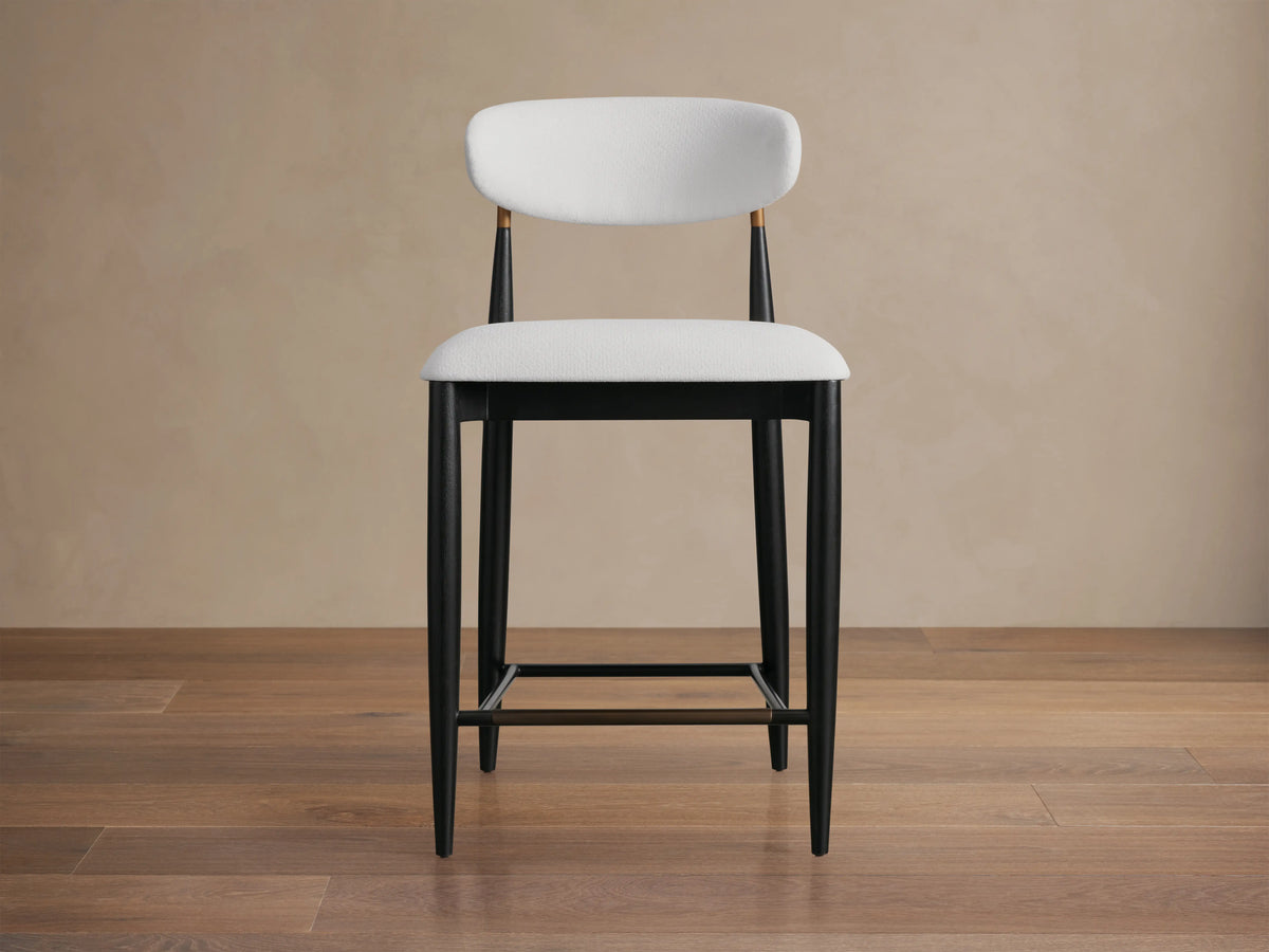 Jagger Armless Stool with sleek black legs, white cushioned seat, and curved backrest supported by bronze bars, on beige background.