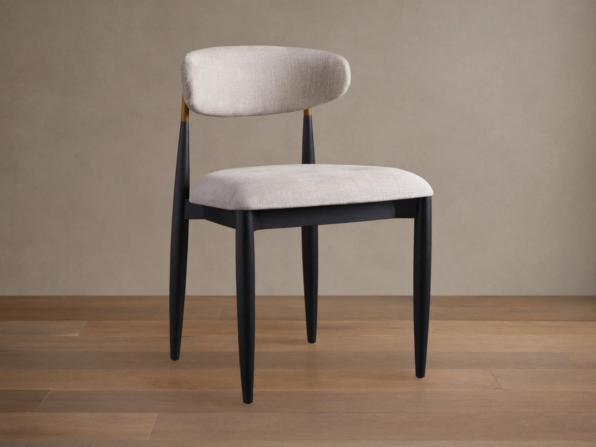 Jagger Dining Side Chair, angled, with black frame, light beige upholstered seat & curved back, gold accent, on wood floor.