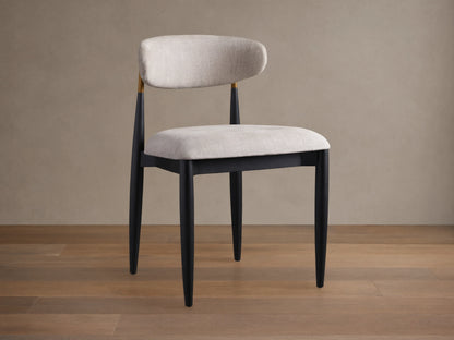 Jagger Dining Side Chair, angled, with black frame, light beige upholstered seat & curved back, gold accent, on wood floor.
