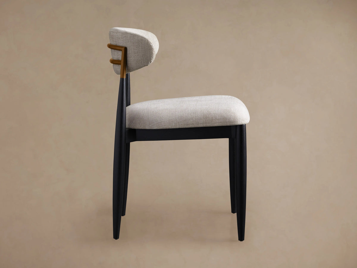 Jagger Dining Side Chair: side view. Light gray upholstered seat & curved back, dark wood frame with golden accent.