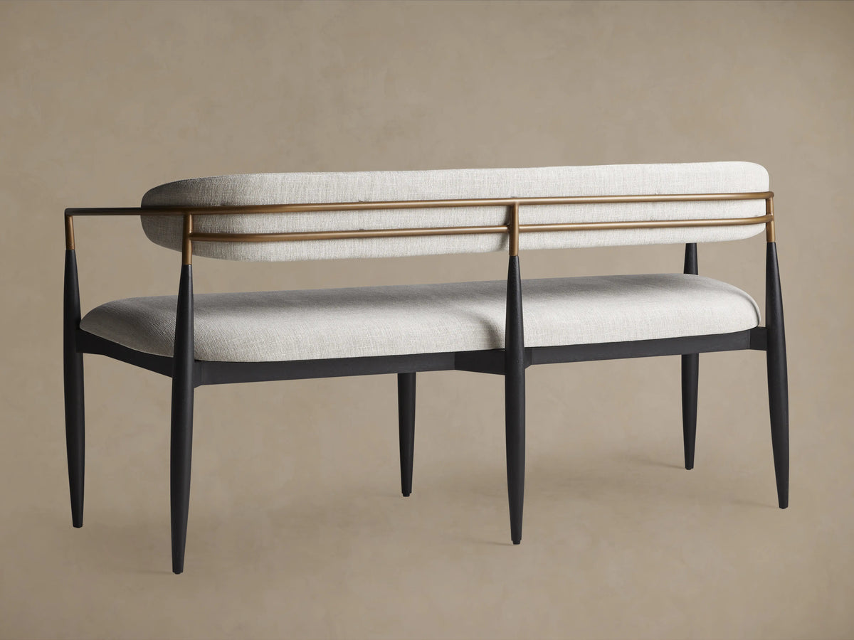 Jagger Dining Bench with soft beige cushions and a dark wood frame. Features slim, pointed legs and a sleek brass bar design.