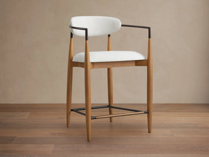 Jagger Stool featuring a white cushioned seat and backrest, wooden legs, and black metal frame, on a wooden floor.