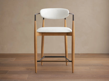 Jagger Stool with a white cushion and backrest, featuring slender wooden legs and black metal accents, set on a wooden floor.