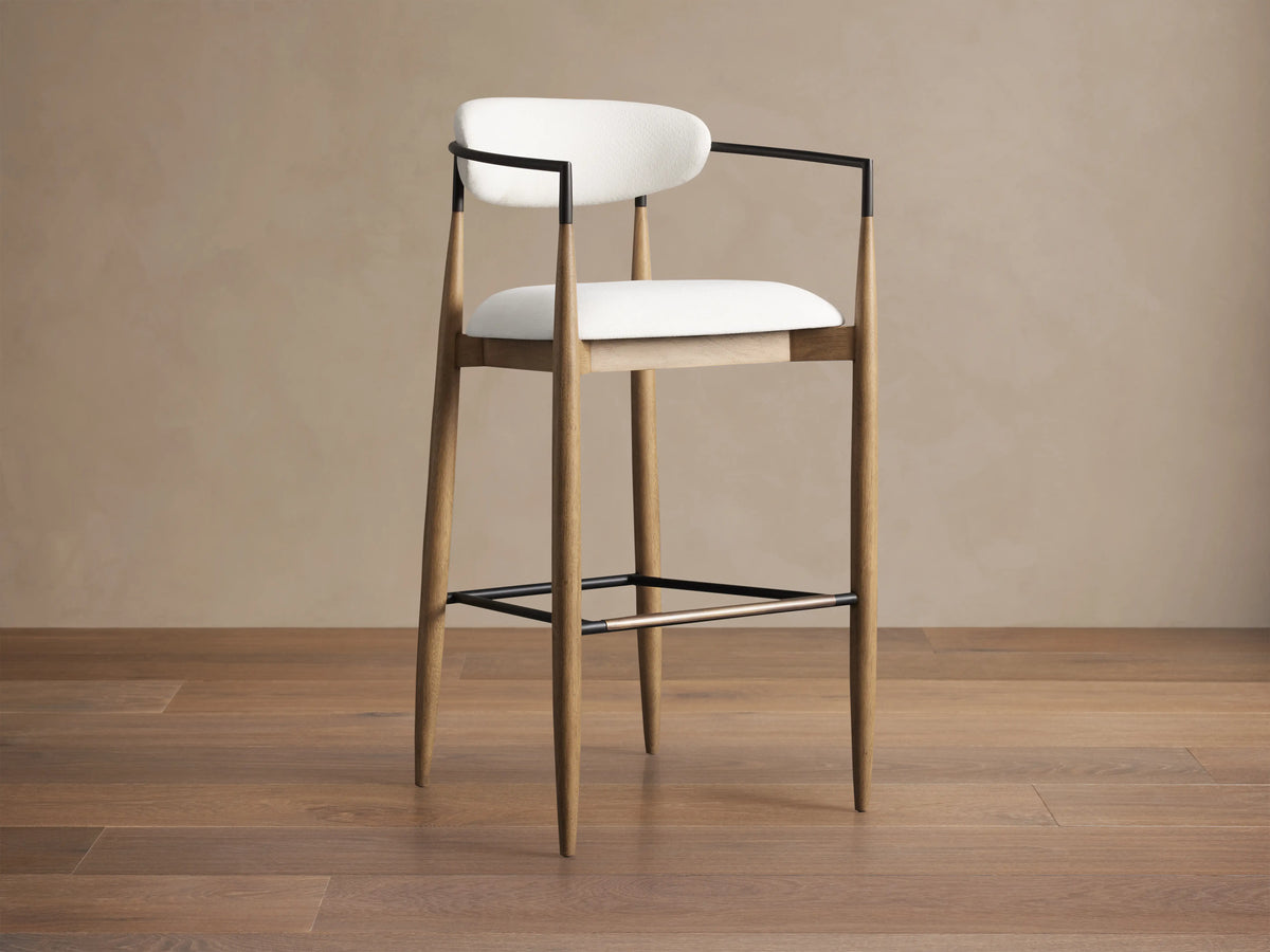 Jagger Barstool with a natural wooden frame, white cushioned seat and backrest, and black metal arms. Placed on wooden floor.