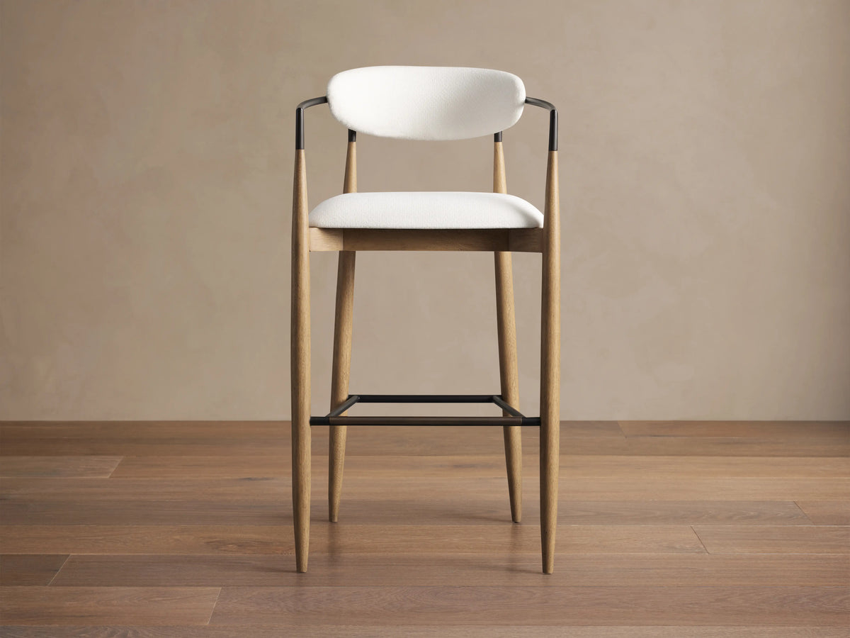 Angled view of Jagger Stool's curved off-white textured performance fabric back, dark bronze frame, and light wood tapered legs.