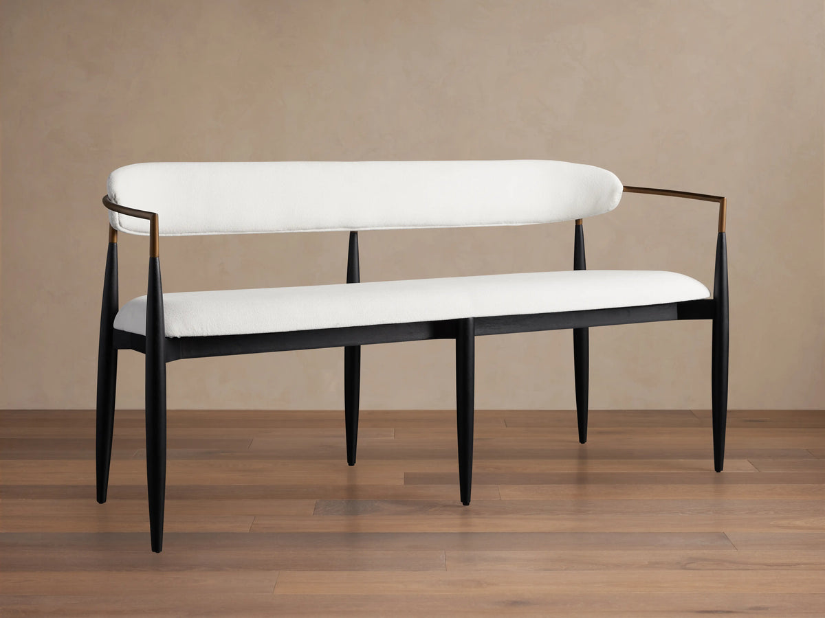 Jagger Dining Bench with white cushioned seat and backrest, black metal frame, on a wooden floor against a beige wall.