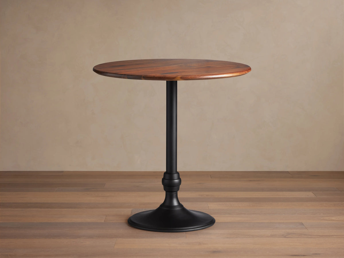 Jacob Bar Table with Wade Base, featuring a round walnut tabletop and a sleek black iron base, set on a wooden floor.
