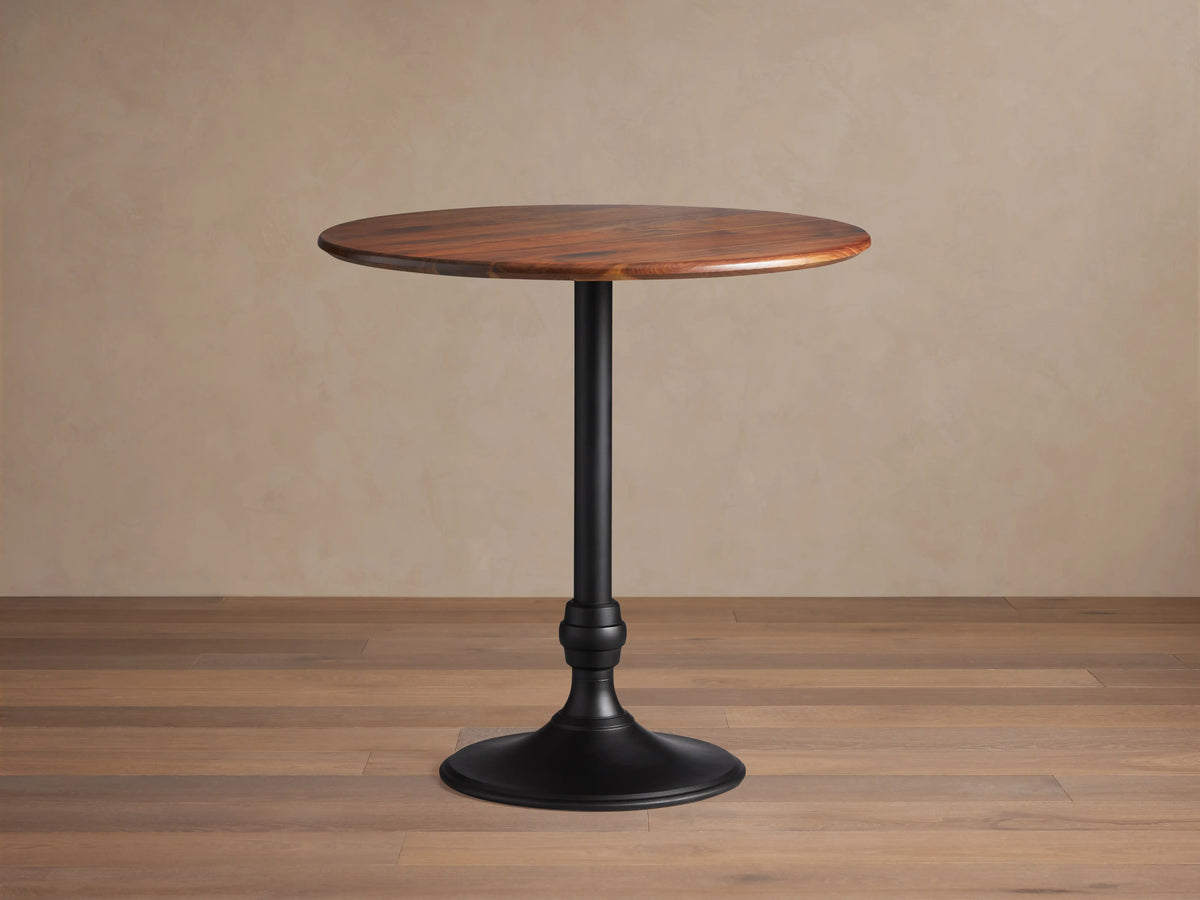Jacob Bar Table with Wade Base, featuring a round walnut tabletop and a sleek black iron base, set on a wooden floor.