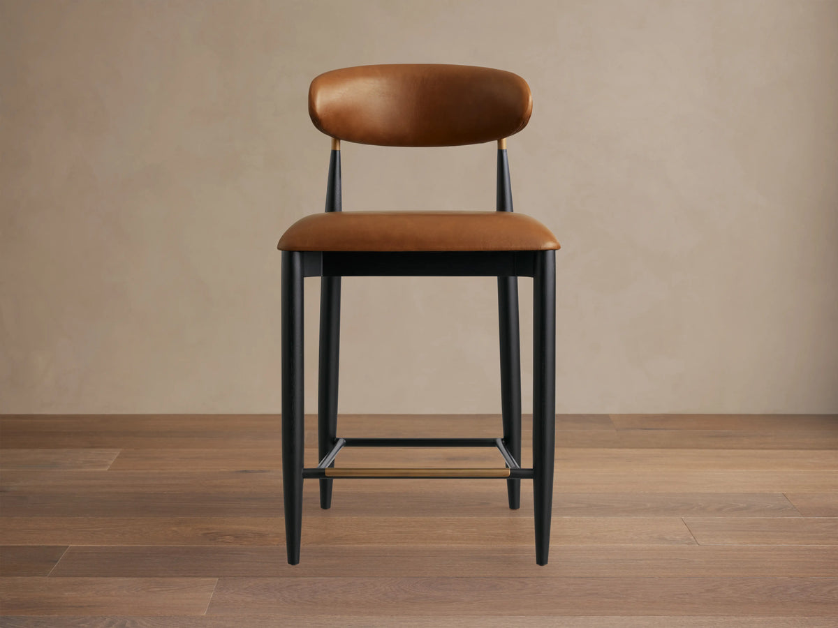 Jagger Faux Leather Armless Stool with brown faux leather seat and back. Black metal frame with gold accents and footrest.