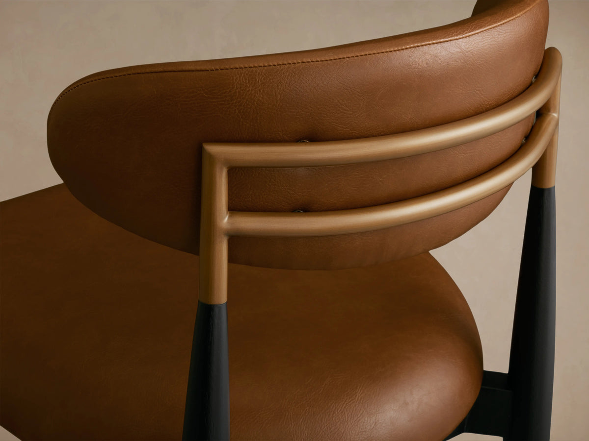 Jagger Faux Leather Armless Stool in tan with a curved backrest and seat. Black metal legs with a footrest on a wooden floor.