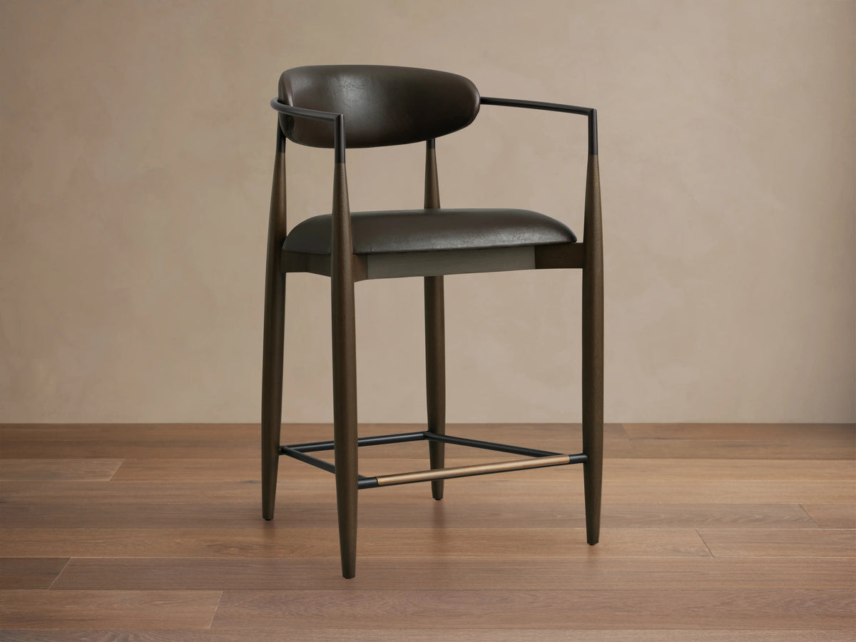 Jagger Faux Leather Stool with dark faux leather seat and backrest, slim metal frame, and wood legs on wooden floor.