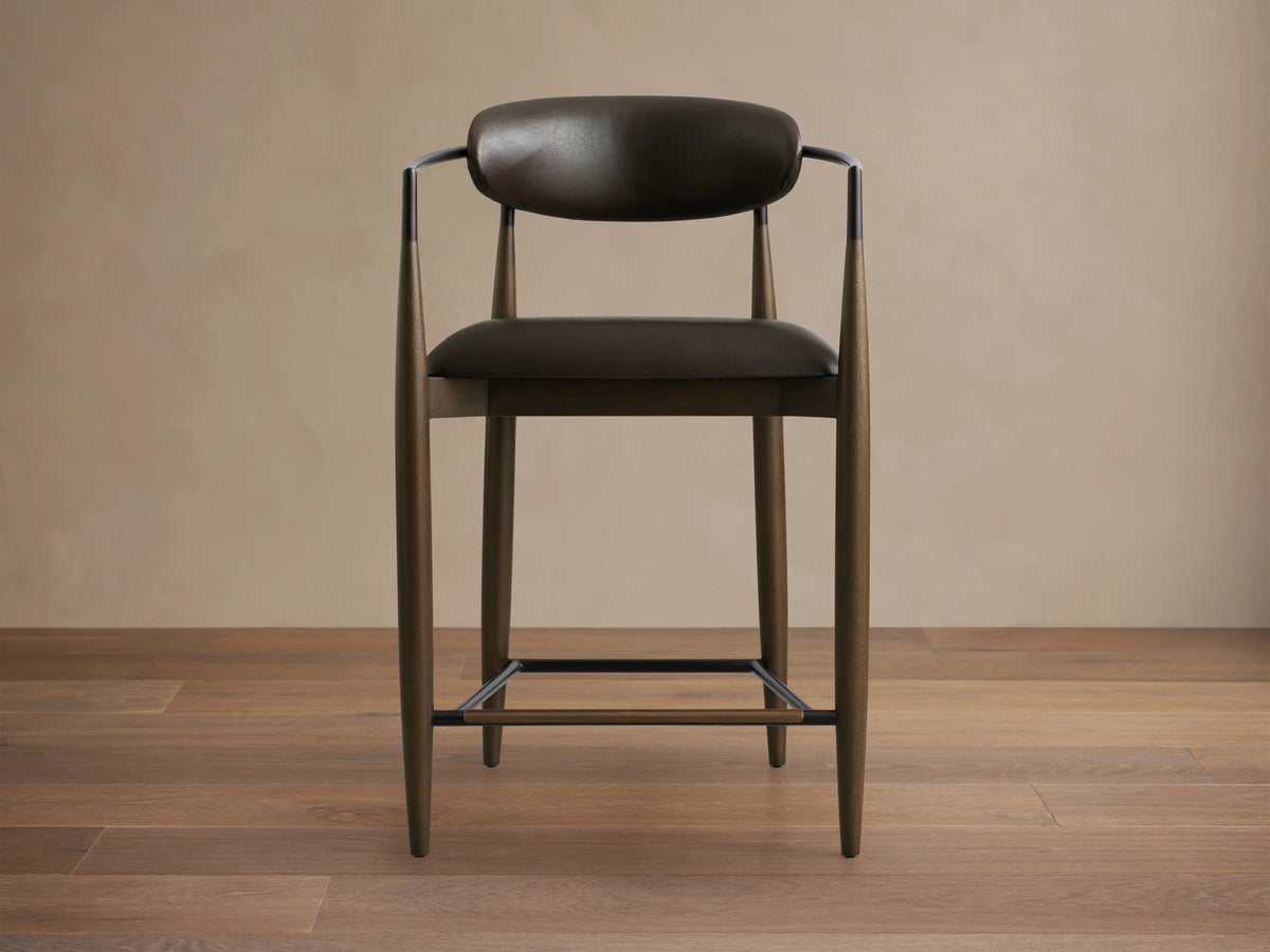 Jagger Faux Leather Stool with dark faux leather seat and backrest, slim metal frame, and wood legs on wooden floor.