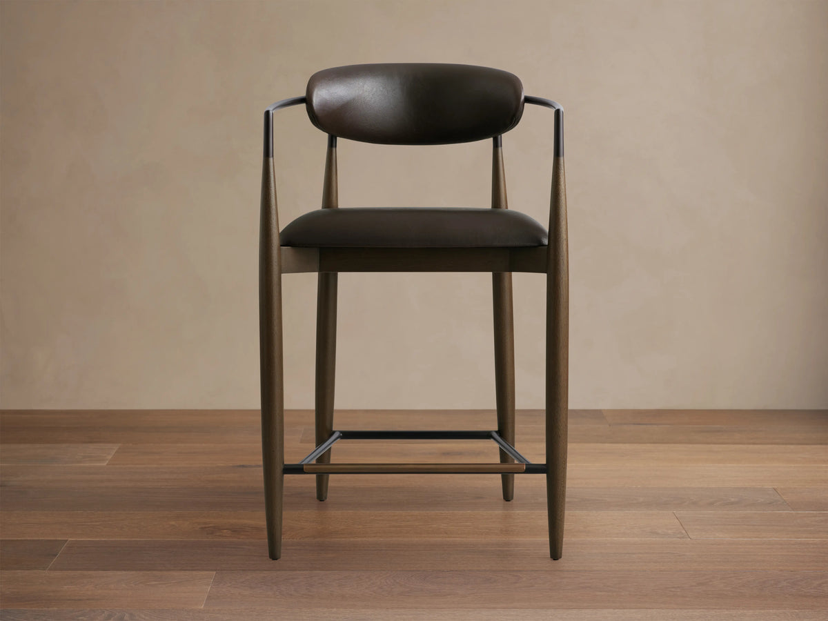 Jagger Faux Leather Stool with dark brown faux leather seat and backrest, slender metal legs, and armrests on a wooden floor.