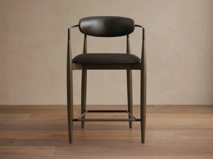 Jagger Faux Leather Stool with dark brown faux leather seat and backrest, slender metal legs, and armrests on a wooden floor.