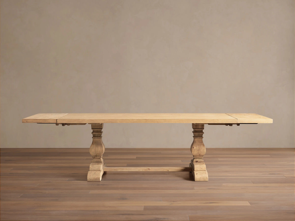 Light natural wood Kensington Dining Table, rectangular top, two chunky carved pedestal bases, shown straight on a wood floor.