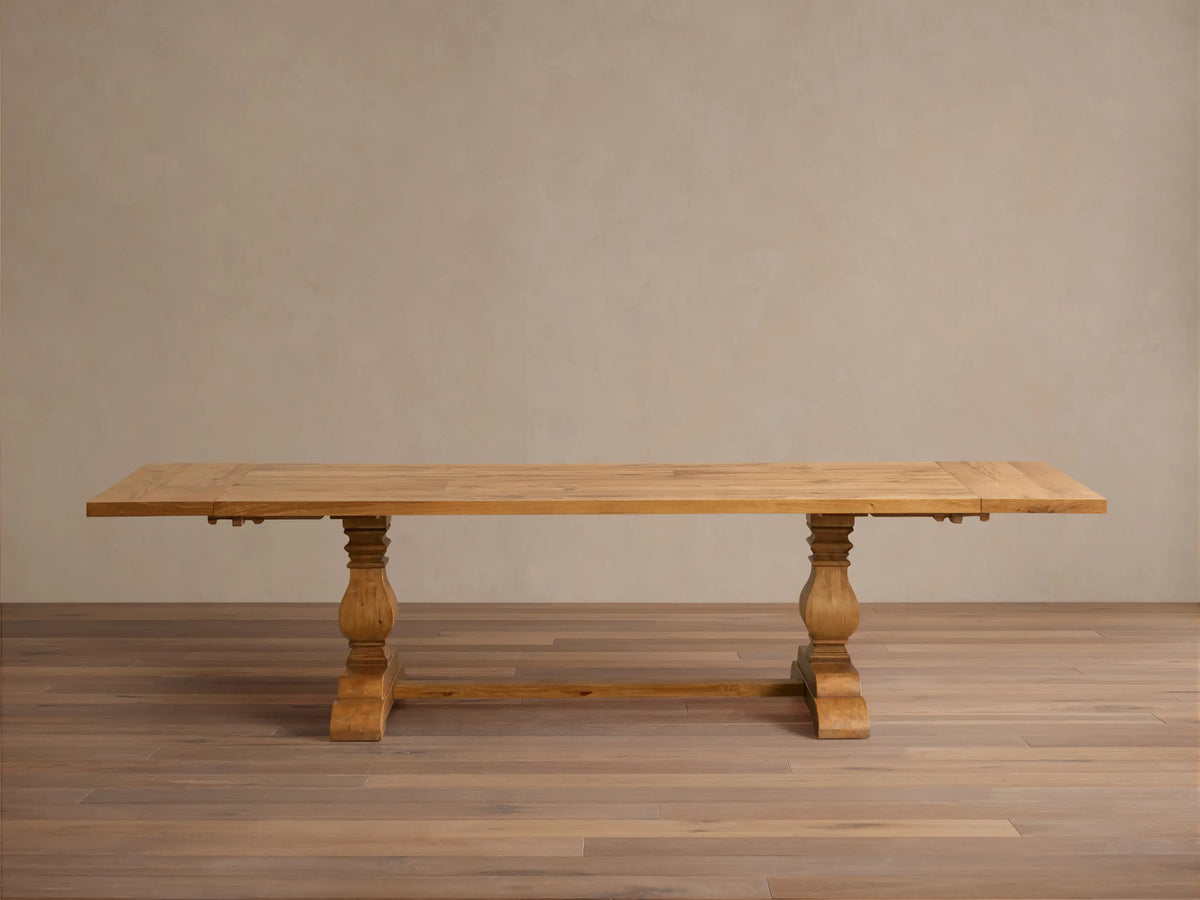 Kensington Reclaimed Dining Table with a light wood rectangular top, supported by two ornate turned pedestal bases, on a wood floor.