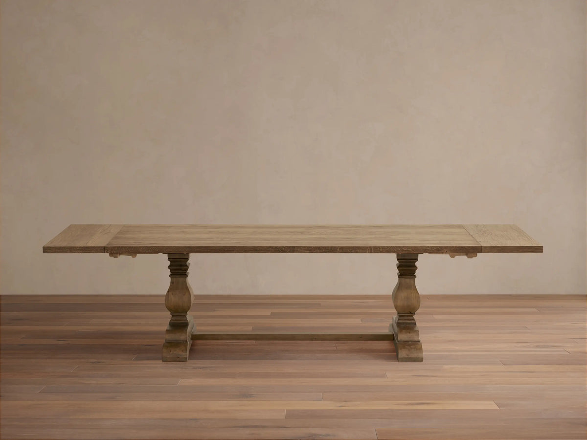 Kensington Reclaimed Dining Table with a rustic wooden top, two intricately carved pedestal legs, on a wooden floor against a neutral background.