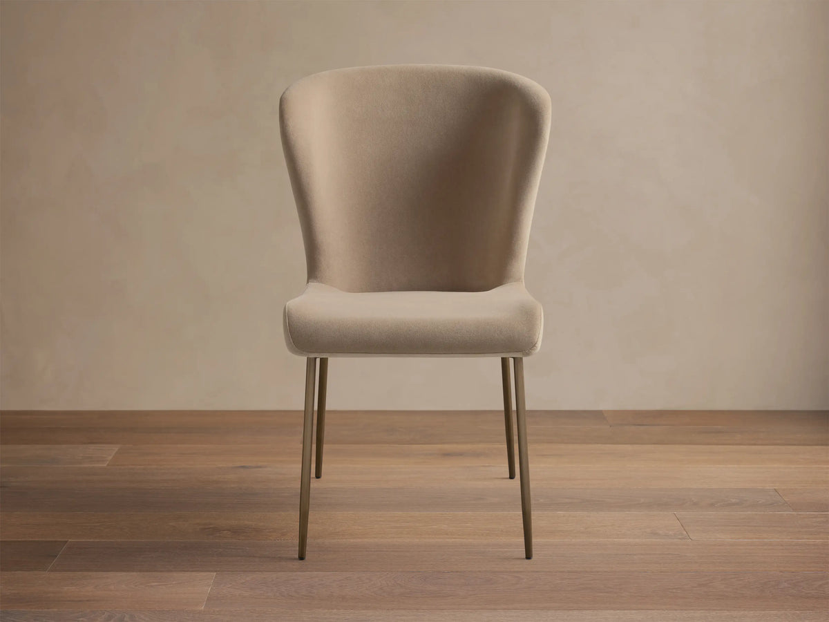 Kirsten High Back Dining Chair, upholstered in light beige, featuring a curved high back and slim metallic bronze legs, angled.