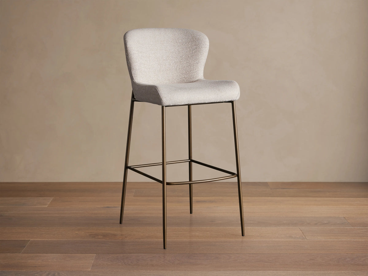Kirsten Stool in Nona Linen with a curved backrest and seat in light fabric, atop slim, powder-coated iron legs on a wooden floor.