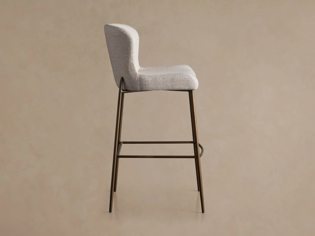 Kirsten Stool in Nona Linen, side view showing light fabric seat and backrest. Brown iron legs with a footrest bar, against a plain background.