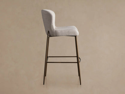 Kirsten Stool in Nona Linen, side view showing light fabric seat and backrest. Brown iron legs with a footrest bar, against a plain background.