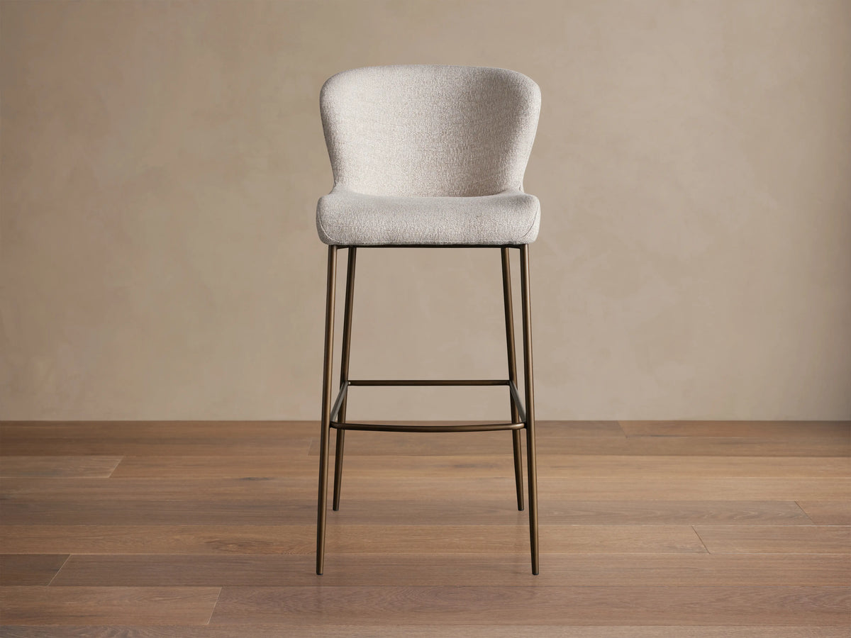 Kirsten Stool in Nona Linen with a curved backrest and seat in light fabric, atop slim, powder-coated iron legs on a wooden floor.