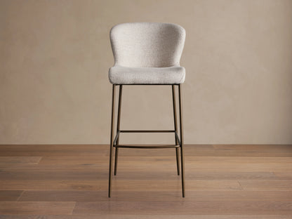 Kirsten Stool in Nona Linen with a curved backrest and seat in light fabric, atop slim, powder-coated iron legs on a wooden floor.