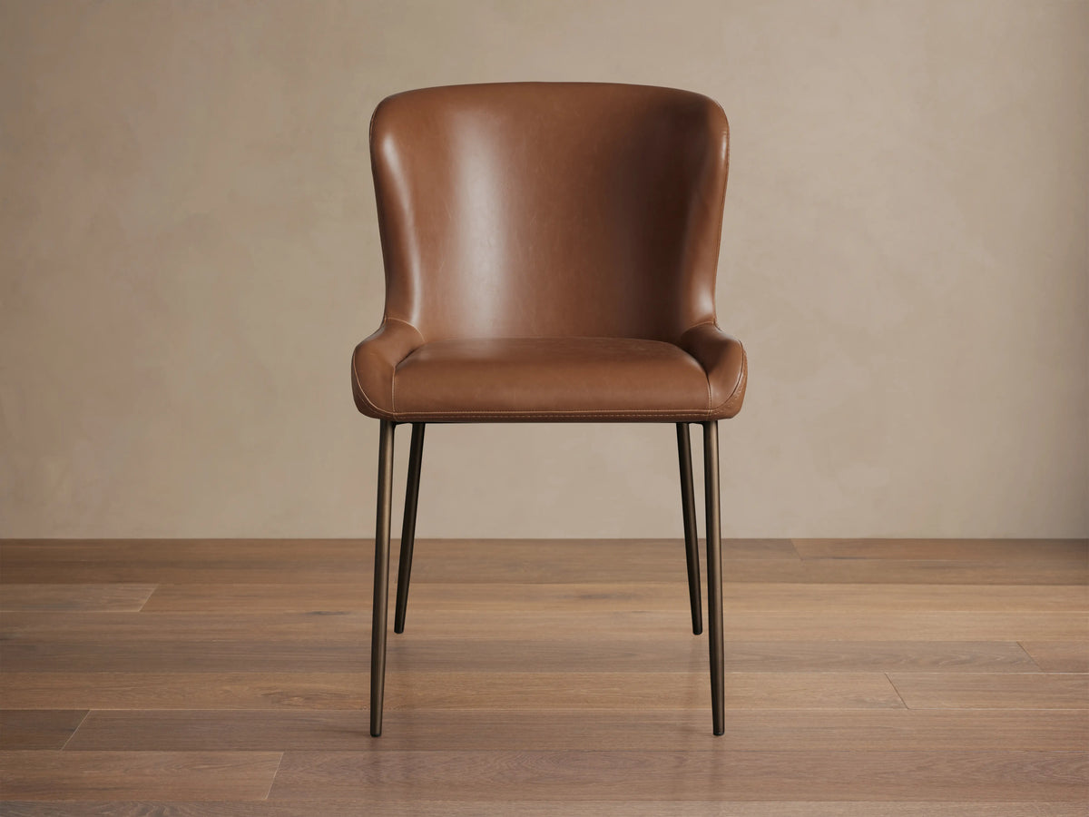 Brown Kirsten Faux Leather Dining Chair with curved backrest and seat. Features slim, black powder-coated iron legs on wooden floor.
