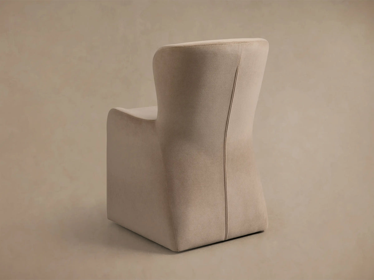 Close-up of the Kirsten Upholstered Dining Chair in soft beige fabric, showing elegant curves of the back and armrest with stitching.