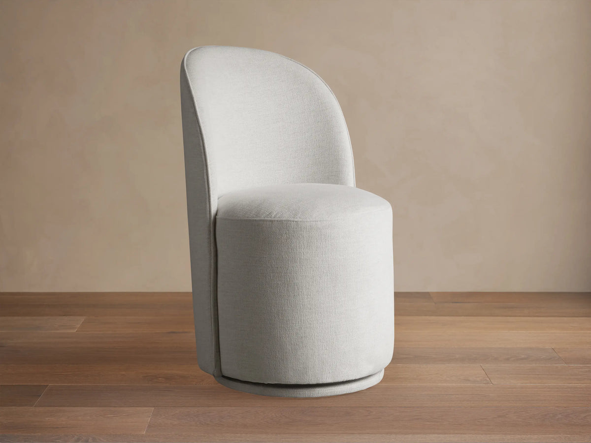 Kira Swivel Dining Chair in Nomad Snow. Light gray fabric, rounded back, cylindrical seat, on wood floor against beige wall.