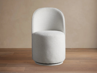 Kira Swivel Dining Chair in Nomad Snow. Light gray fabric, rounded back, cylindrical seat, on wood floor against beige wall.
