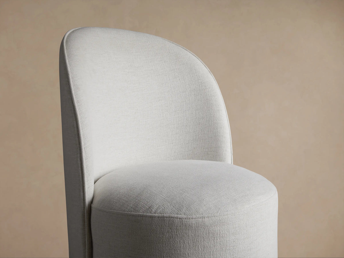 Kira Swivel Dining Chair in Nomad Snow. Light grey, curved fabric chair with a round swivel base, centered against a warm wall.