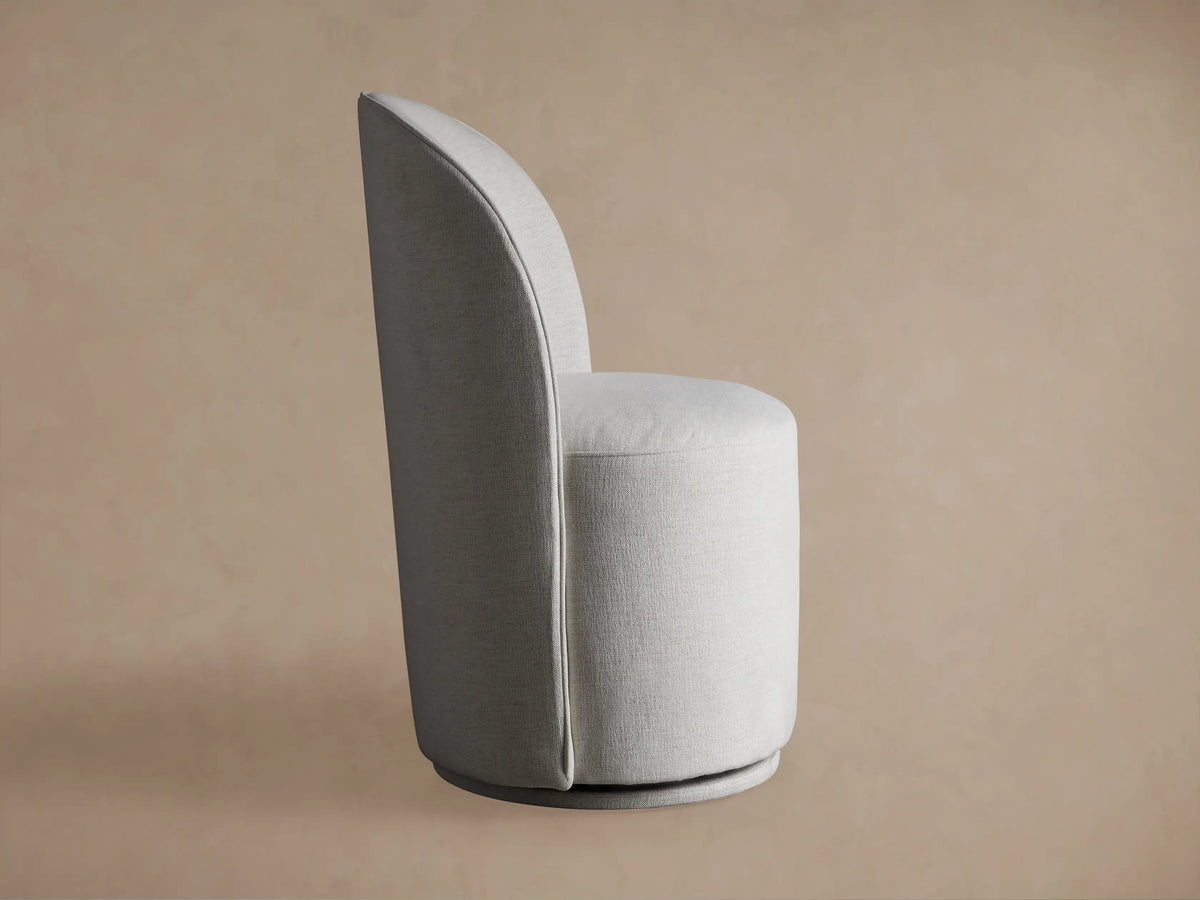 Kira Swivel Dining Chair in Nomad Snow. Close-up shows curved back, round seat, light off-white textured fabric against tan background.