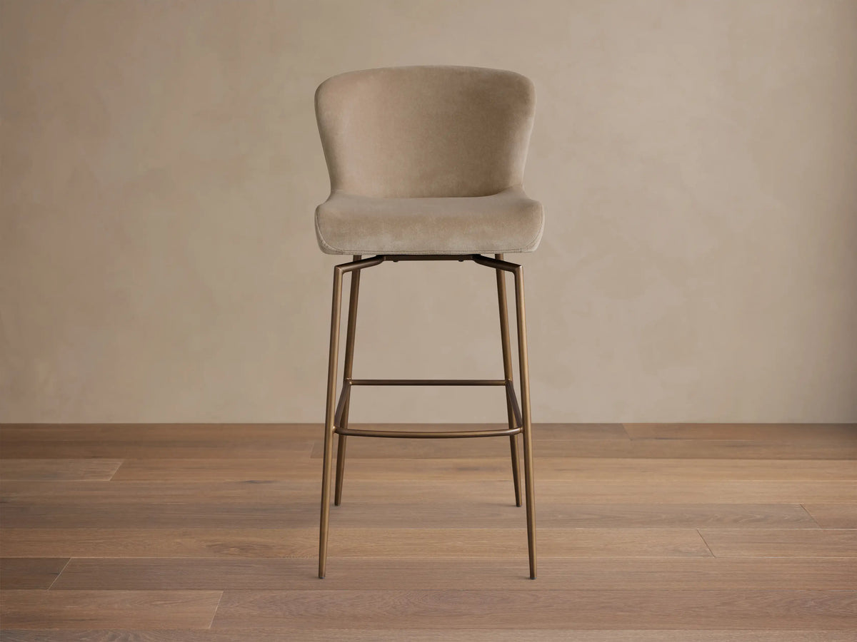 Front view of the beige upholstered Kirsten Swivel Stool with a curved back and slender bronze iron legs.