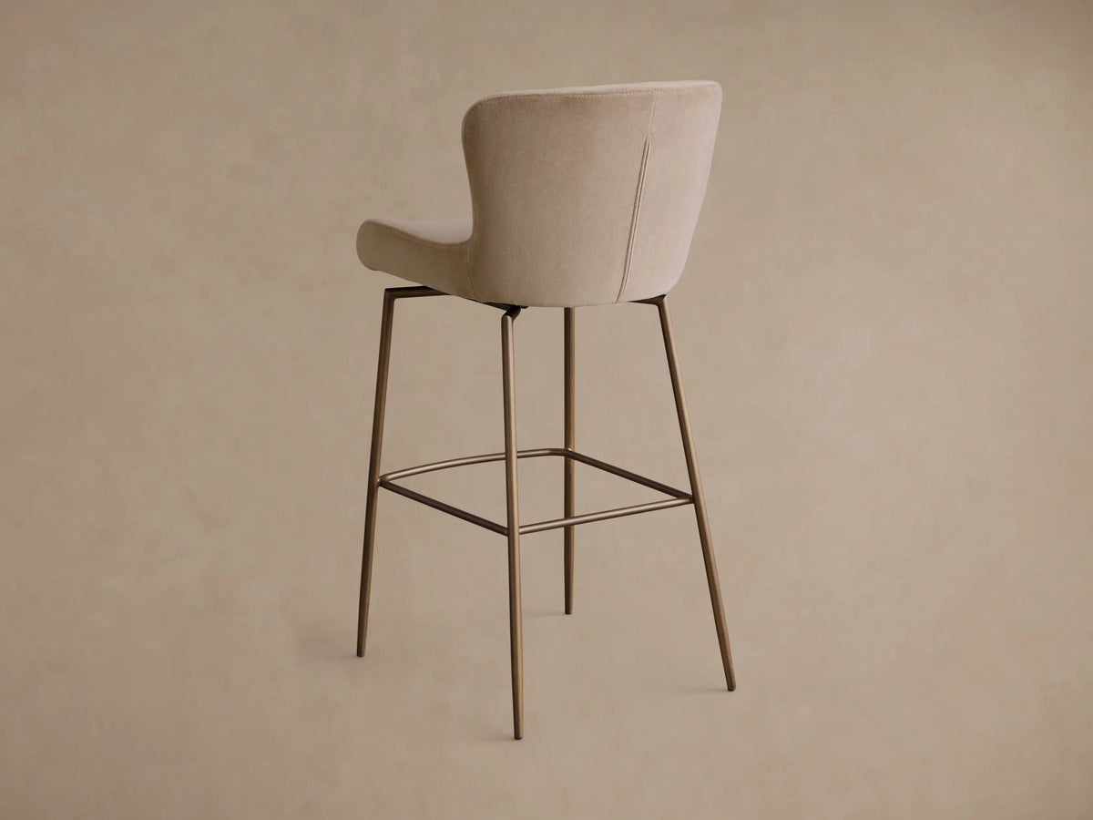Close-up of the beige Kirsten Swivel Stool showing its curved upholstered seat and backrest with brown metal frame.