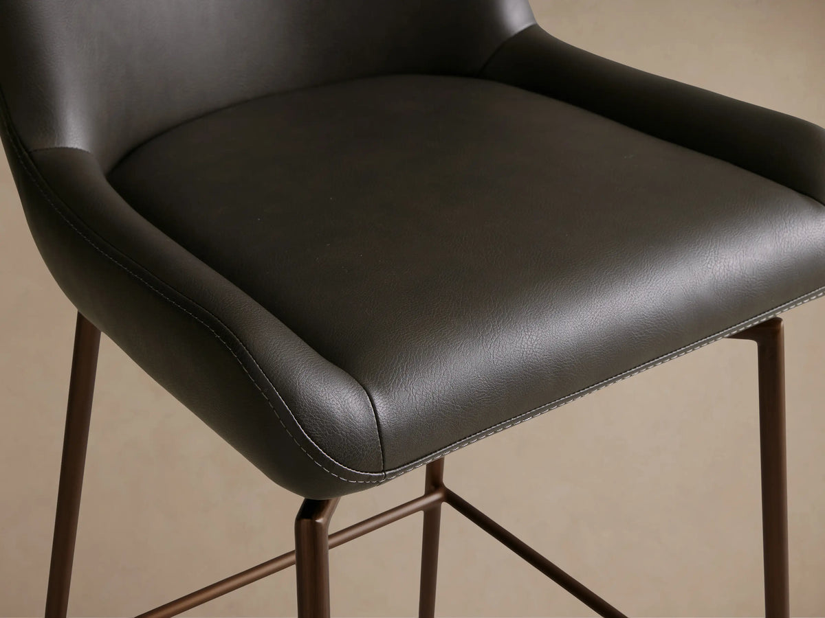 Kirsten Faux Leather Swivel Stool in dark faux leather with a curved backrest and seat, metal legs, on wooden floor.