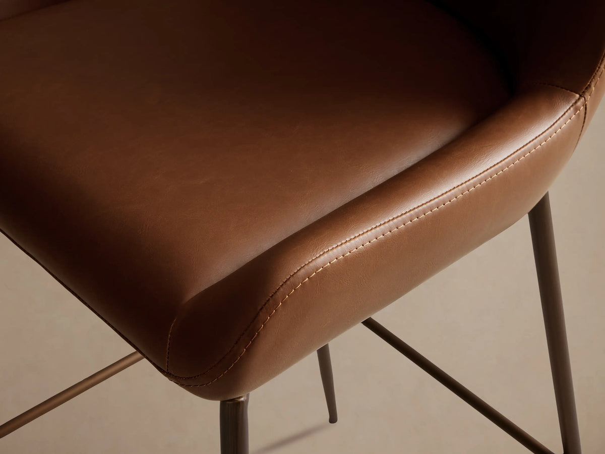 Kirsten faux leather swivel stool in brown, with a curved backrest and seat. Supported by a powder-coated iron frame and legs.