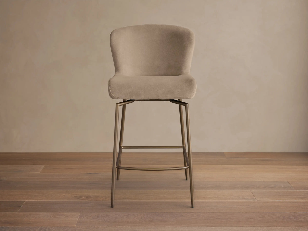 Front view of a light beige upholstered Kirsten Swivel Stool with a curved back and bronze metal legs and footrest.
