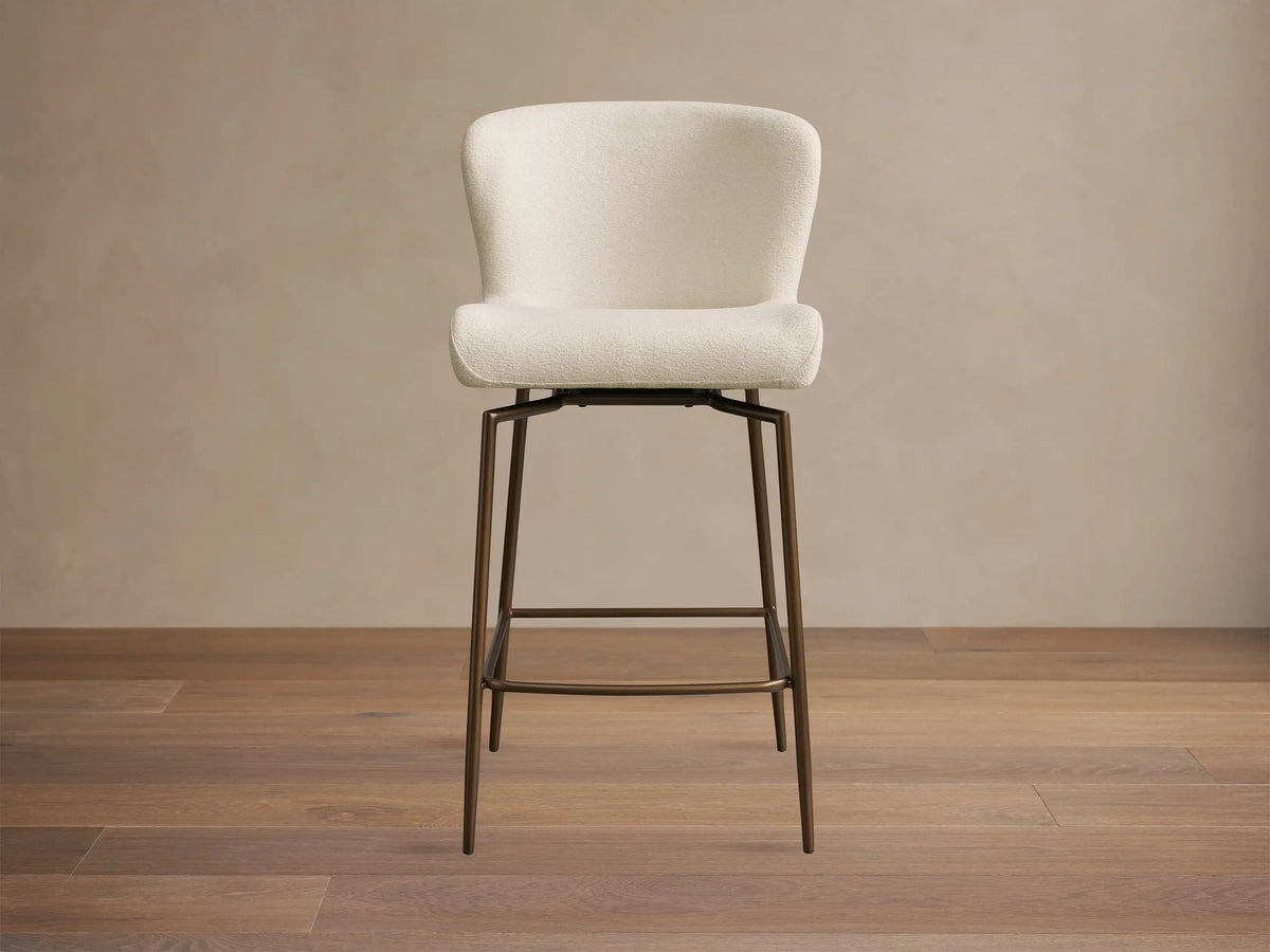 Kirsten Swivel Stool with cream upholstered curved back and seat, tall dark bronze iron legs, and footrest on a wooden floor.